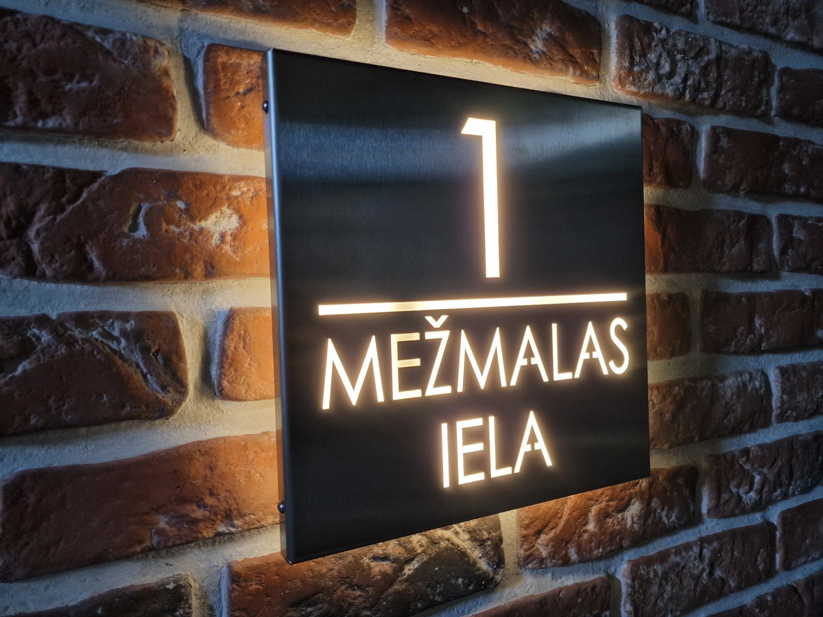 Stainless-steel square LumiSign with illuminated number 1 and two-line MEŽMALAS IELA lettering on a brick wall.